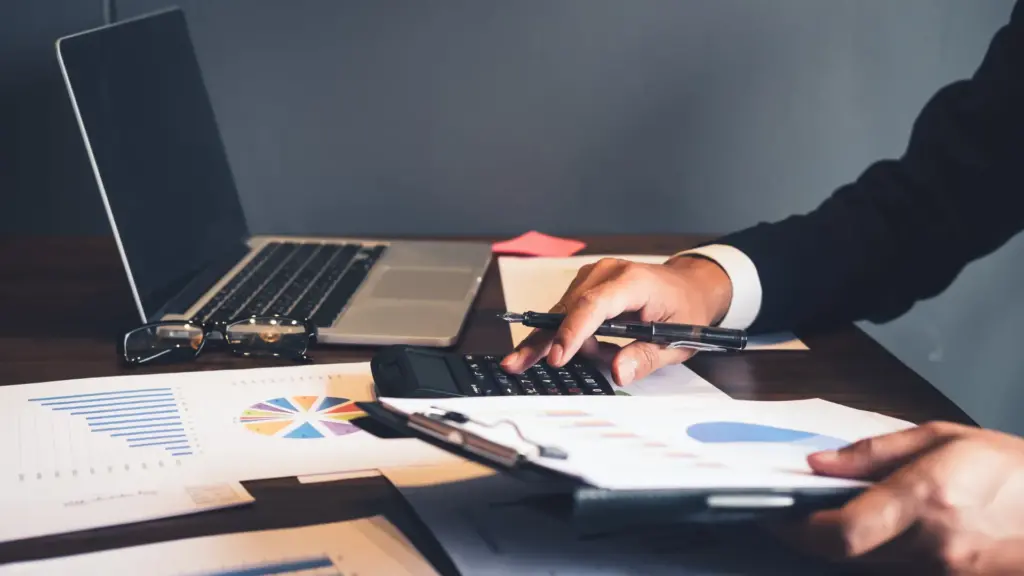 Person using a calculator and reviewing financial charts and graphs on a desk with a laptop and documents.