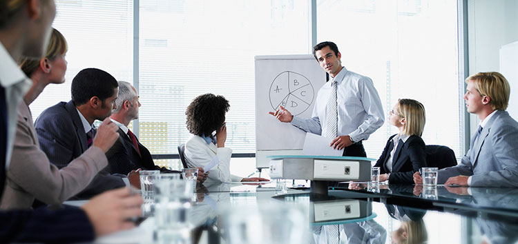 A man presents a pie chart on a flip chart to a group of colleagues in a conference room. Attendees are seated around a table with documents and glasses of water.