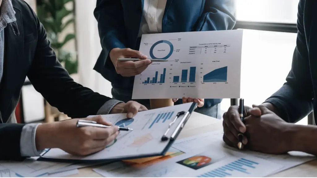 Three people analyzing printed bar and pie charts in a meeting, with documents and graphs on the table.