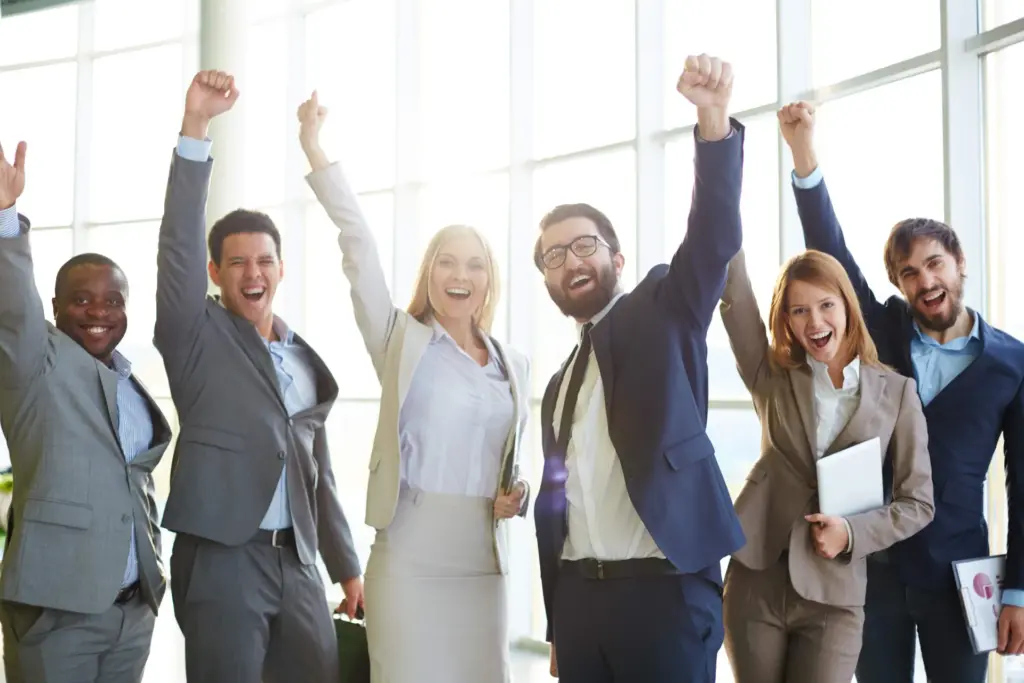Group of six people in business attire standing indoors, raising their arms in celebration, and smiling at the camera.