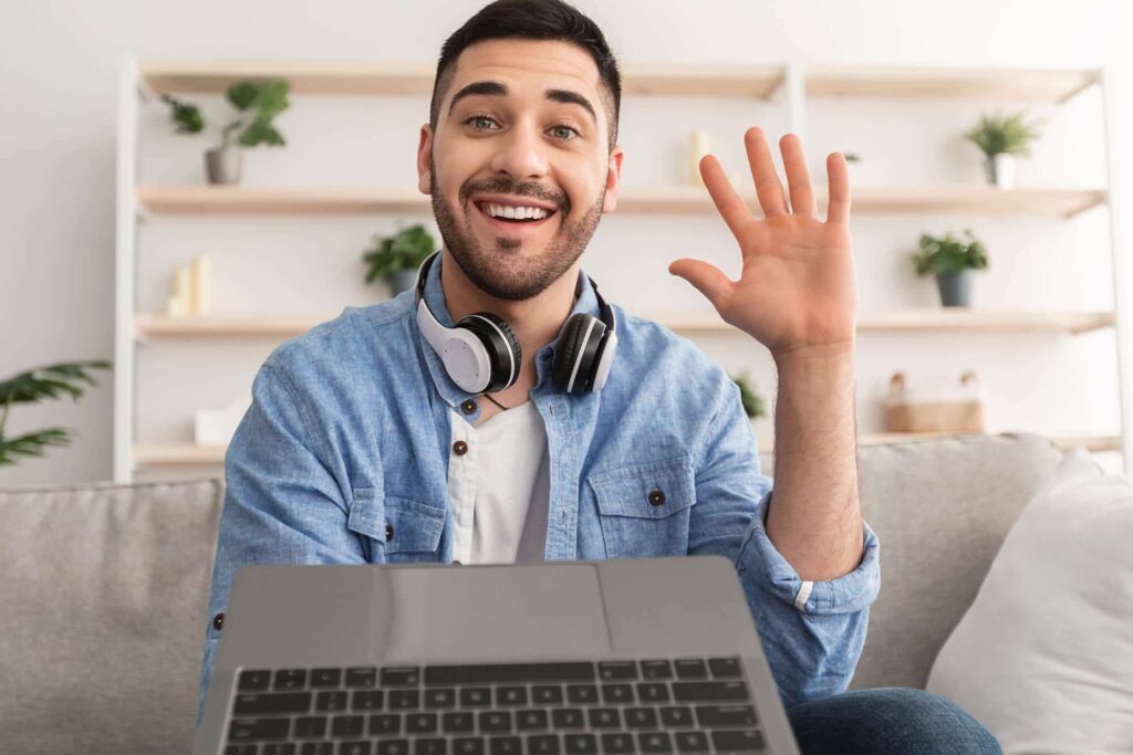 Smiling person with headphones waves at the camera, sitting with a laptop. Shelves with plants are visible in the background.