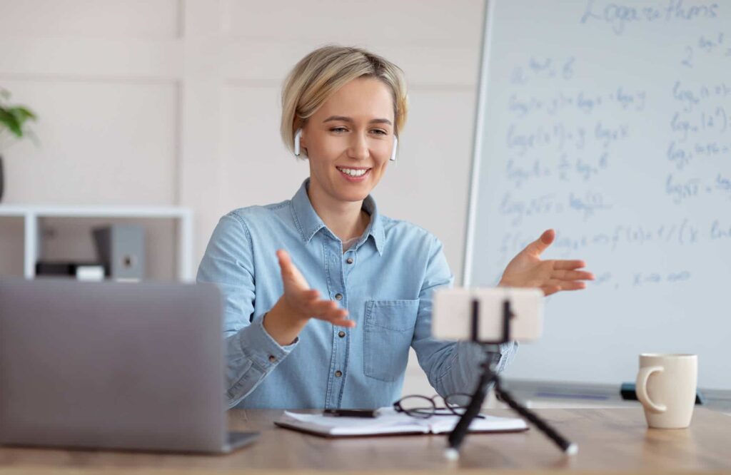 Person with earbuds gestures during a video call at a desk with a laptop, smartphone on a tripod, notebook, and mug. Whiteboard with writing in the background.