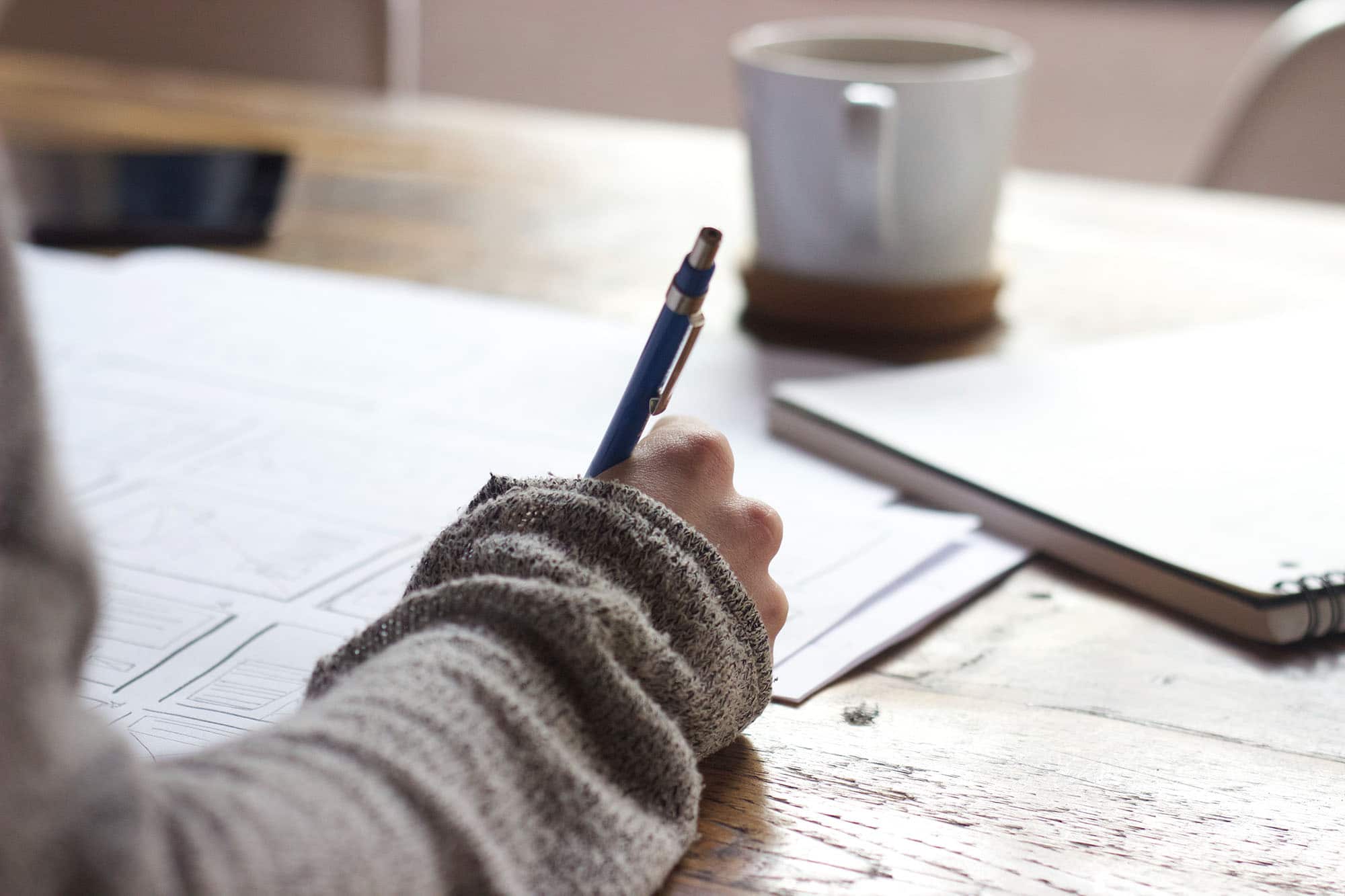 A person in a gray sweater writes on papers at a wooden table, with a notebook, a smartphone, and a coffee cup nearby.