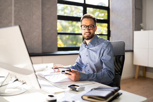 A man in glasses sits at a desk with papers, a pen, and a smartphone. He works on a computer in a bright office with large windows. He is smiling.