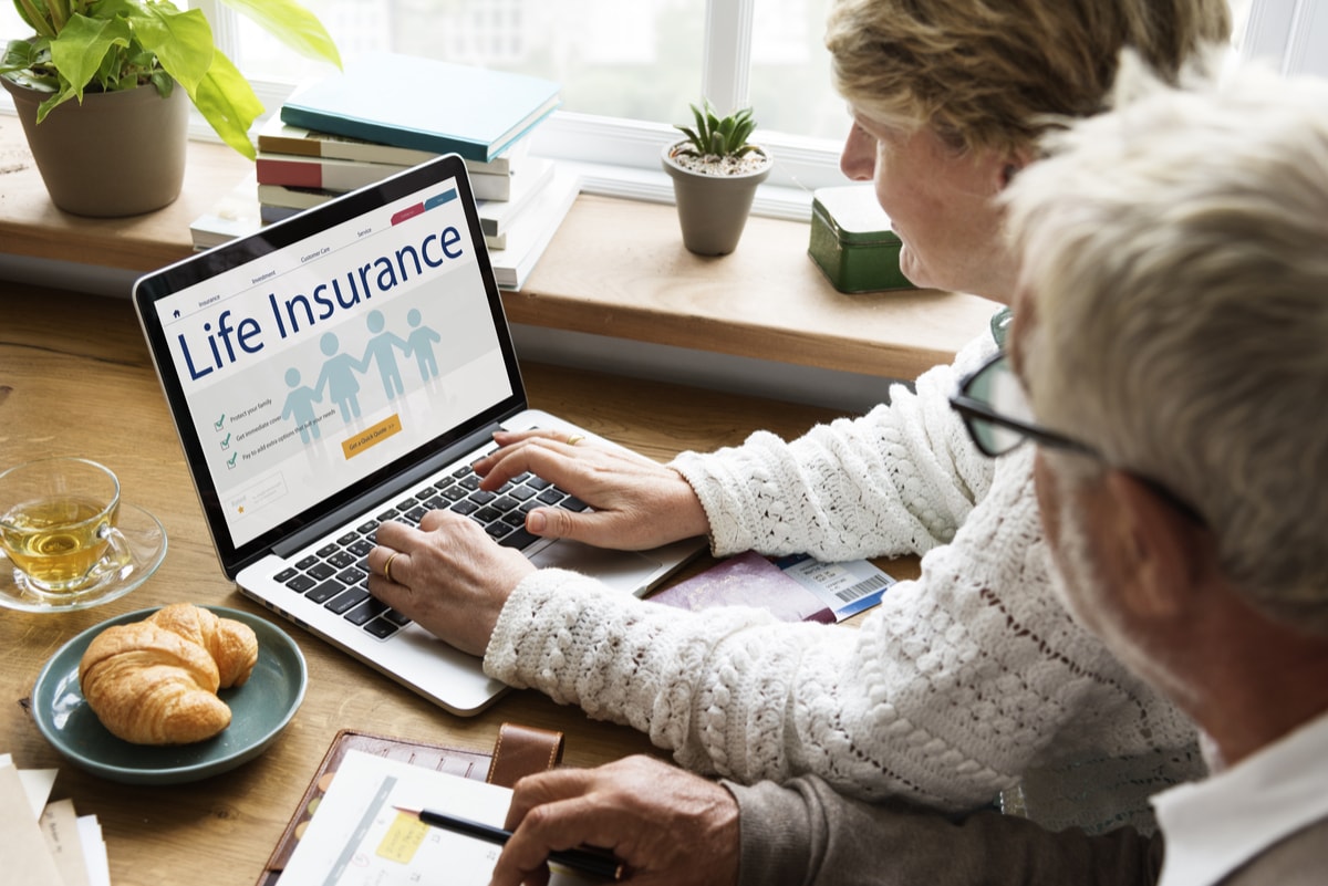 Two people sit at a table with a laptop displaying a life insurance webpage. Croissants, plants, and books are on the table.