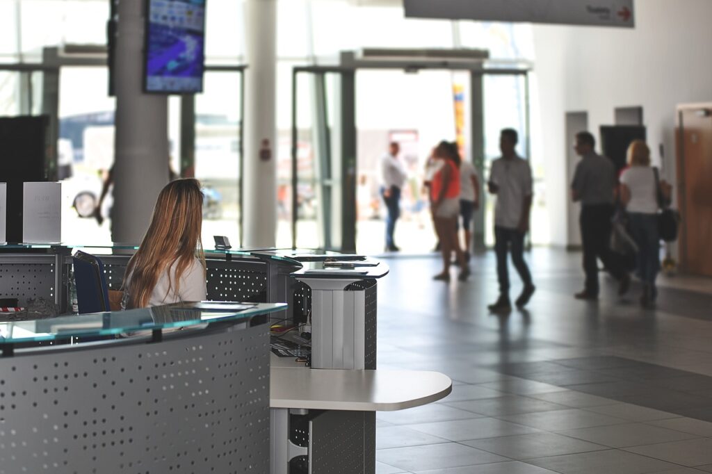 A woman with long hair sits at a modern reception desk in a spacious building lobby. People walk in the background near large glass doors.