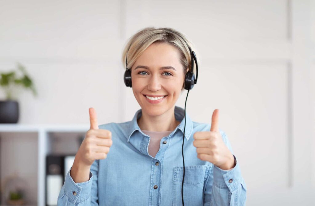 Person with a headset smiling and giving two thumbs up, wearing a light blue shirt indoors with a plant in the background.