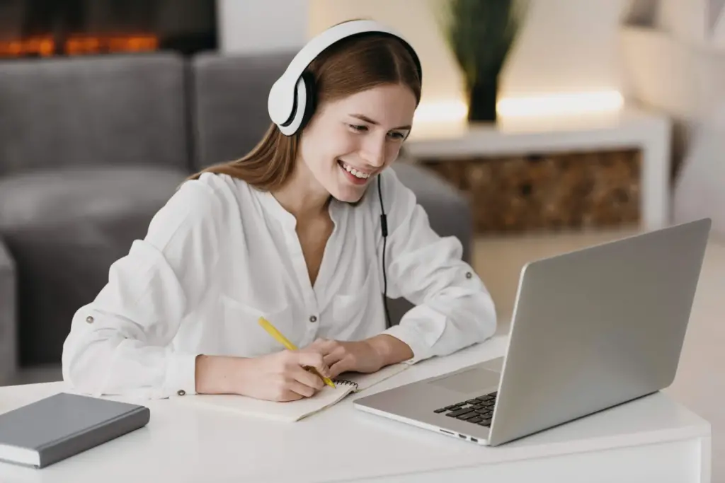 Person wearing headphones sits at a desk, smiling and writing in a notebook, while looking at a laptop.