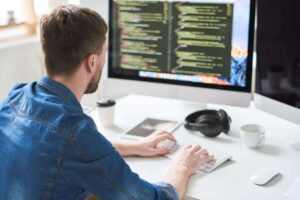 A person in a blue shirt types code on a keyboard, facing a computer screen displaying colorful programming text. A coffee cup, headphones, and a notebook are on the white desk.