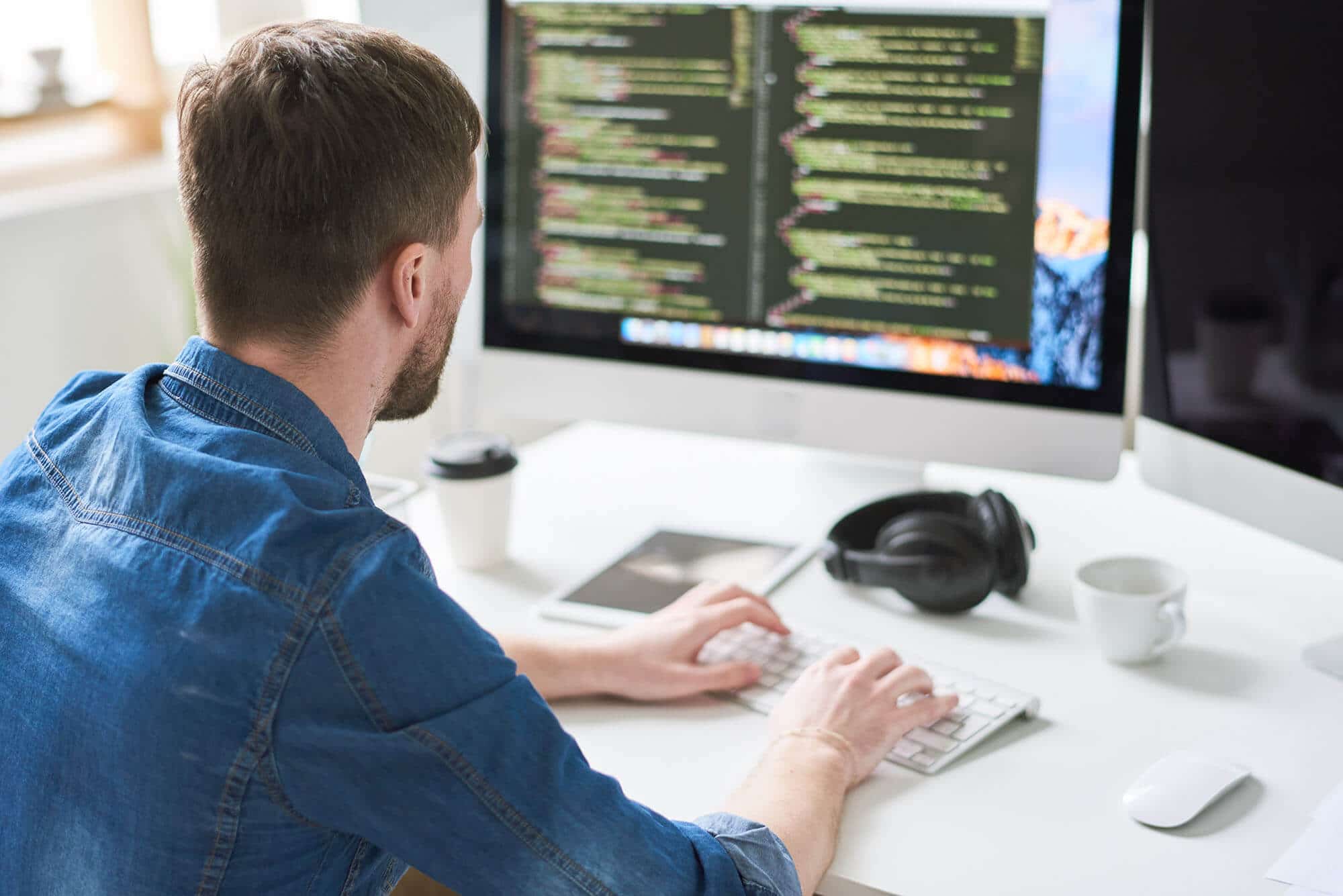A person in a blue shirt types code on a keyboard, facing a computer screen displaying colorful programming text. A coffee cup, headphones, and a notebook are on the white desk.