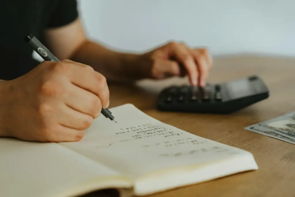 Hands holding a pen and using a calculator on a wooden table. Nearby is an open notebook with calculations and a hundred-dollar bill partially visible.