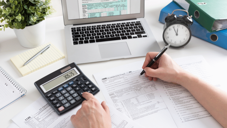 Person using a calculator and filling out tax forms at a desk with a laptop, documents, clock, folders, notepad, and a small potted plant.