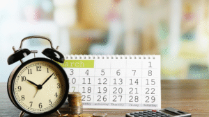 A black alarm clock, a desk calendar open to March, stacks of coins, and a calculator on a wooden table.