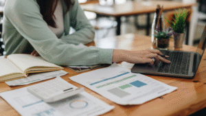Person working at a desk with a laptop, open notebook, calculator, smartphone, and financial documents with charts and graphs.