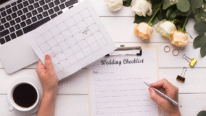 Person holding a calendar with a heart on one date, writing on a wedding checklist by a laptop, coffee, roses, and wedding rings.