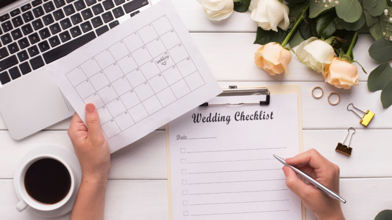 Person holding a calendar with a heart on one date, writing on a wedding checklist by a laptop, coffee, roses, and wedding rings.